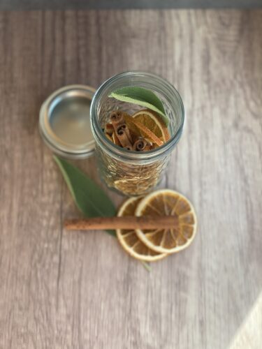 A clear glass jar filled with dried orange slices, cinnamon sticks, and fresh sage leaves, with a lid beside it and additional orange slices on the wooden surface.