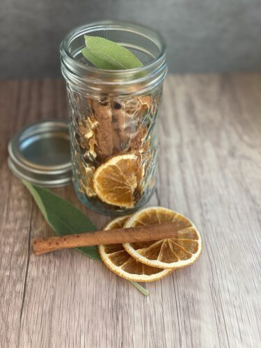 A glass jar filled with dried orange slices, cinnamon sticks, and sage leaves, placed on a wooden surface.