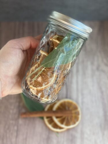 A hand holding a glass jar filled with dried orange slices, fresh sage leaves, and wrapped in clear plastic, with cinnamon sticks placed beside it.