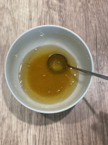 A white bowl containing a mixture of melted beeswax and honey, with a silver spoon resting inside, on a wooden surface.