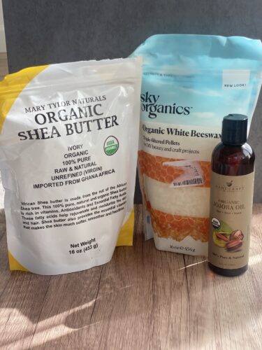 An assortment of organic skincare ingredients, including a bag of organic shea butter, a bag of white beeswax pellets, and a bottle of jojoba oil, displayed on a wooden surface.