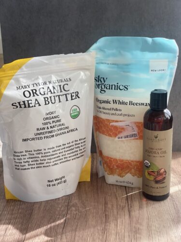 A package of organic shea butter, a bag of organic white beeswax pellets, and a bottle of jojoba oil displayed on a wooden surface.