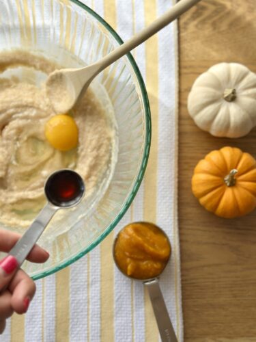A hand adding vanilla extract to a bowl of batter, with a spoon and small pumpkins nearby, on a striped yellow and white kitchen towel.