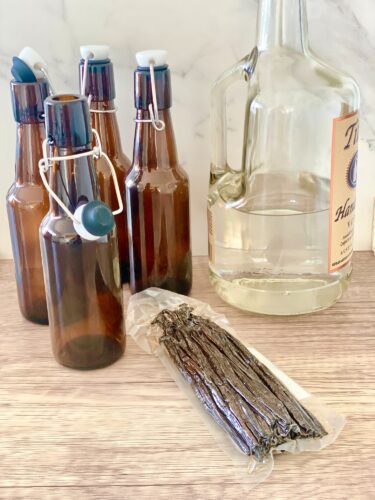 A collection of amber glass bottles with swing top caps next to a large bottle of alcohol and several whole vanilla beans on a wooden surface.