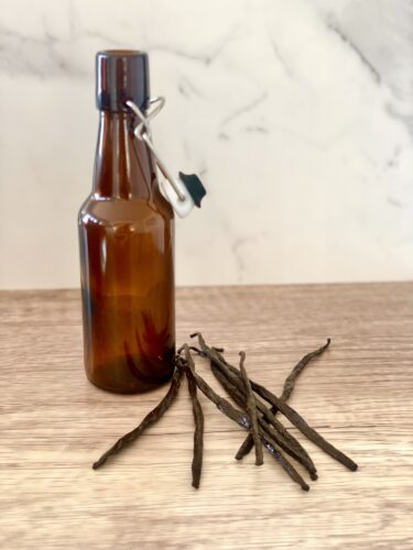 An amber glass bottle with a swing top, next to several whole vanilla beans, placed on a wooden surface against a light-colored background.