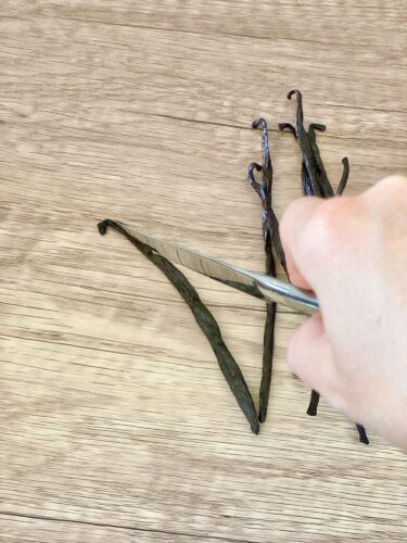 A hand holding a knife, slicing open organic vanilla beans on a wooden surface.