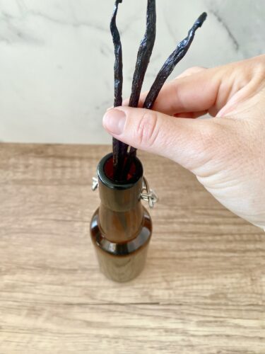 A hand inserting vanilla beans into a swing-top glass bottle on a wooden surface.