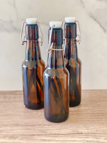 Three amber glass bottles with swing-top lids, each containing vanilla beans, placed on a wooden surface.