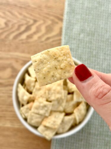 A hand holding a homemade dill cracker above a bowl filled with more crackers, showcasing the snack's texture and color.