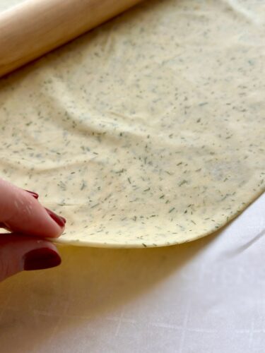 A close-up of rolled-out cracker dough sprinkled with dill, held by a hand with red nail polish, next to a wooden rolling pin.
