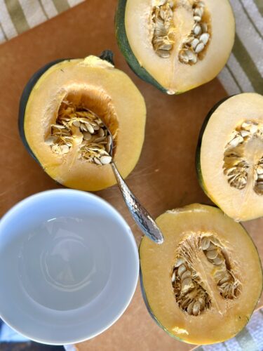 Halved acorn squash with seeds exposed and a spoon resting beside a small bowl of water on a wooden cutting board.
