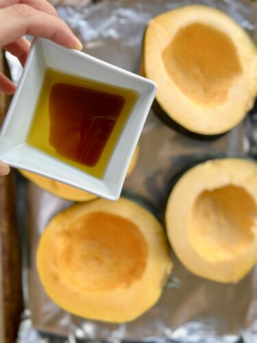 A hand holding a small white bowl of olive oil above halved acorn squash pieces resting on a baking sheet covered with aluminum foil.