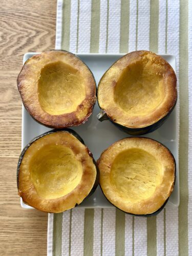 Four roasted acorn squash halves placed cut side up on a white plate with a striped tablecloth underneath.