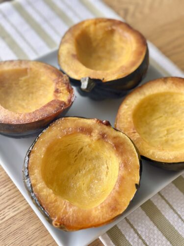 Four halved acorn squashes, perfectly roasted, displayed on a white plate with a light wooden background.