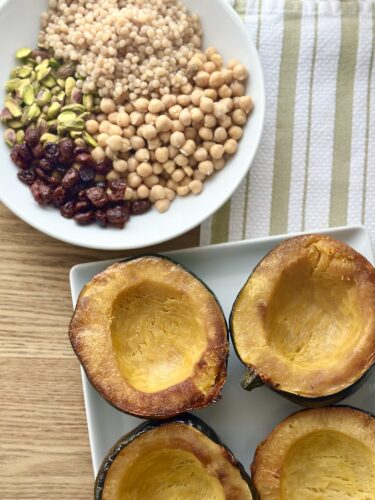 A plate featuring roasted acorn squash halves next to a bowl of cous cous, pistachios, chickpeas, and dried cranberries, highlighting the ingredients for a stuffed acorn squash recipe.