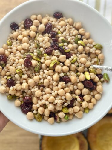A bowl filled with a colorful mixture of cous cous, chickpeas, dried cranberries, and pistachios, with a spoon resting inside, showcasing the texture and ingredients.