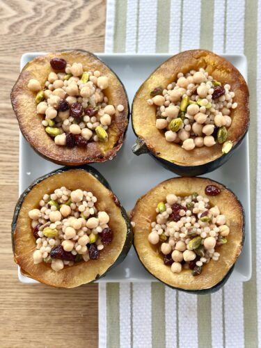 Four halved acorn squash filled with a mixture of couscous, chickpeas, cranberries, and pistachios, arranged on a plate.