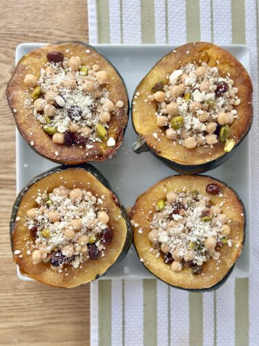 Four halves of roasted acorn squash filled with a mixture of cous cous, chickpeas, pistachios, cranberries, and crumbled feta, arranged on a white plate with a striped fabric background.
