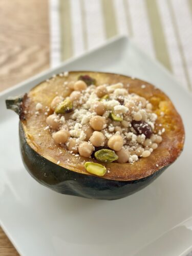 Stuffed acorn squash filled with pumpkin pie spice cous cous, chickpeas, pistachios, cranberries, and crumbled feta on a white plate.