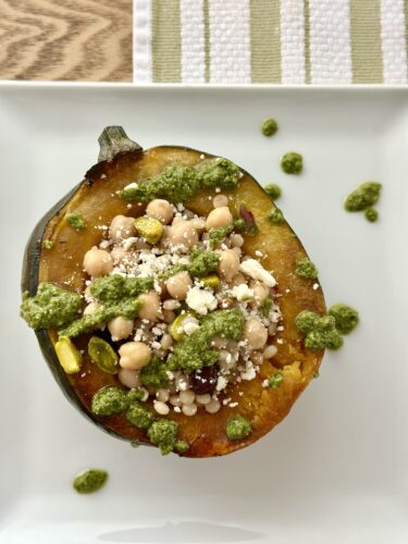 A roasted acorn squash half filled with pumpkin pie spice cous cous, chickpeas, dried cranberries, and pistachios, topped with crumbled feta cheese and a drizzle of green pesto on a white plate.