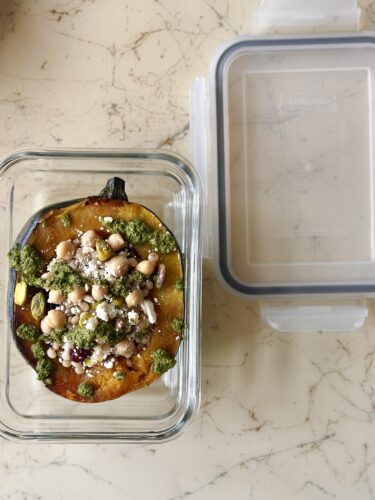 Stuffed acorn squash with cous cous, pistachios, cranberries, chickpeas, and feta in a glass container with a transparent lid on a marble surface.