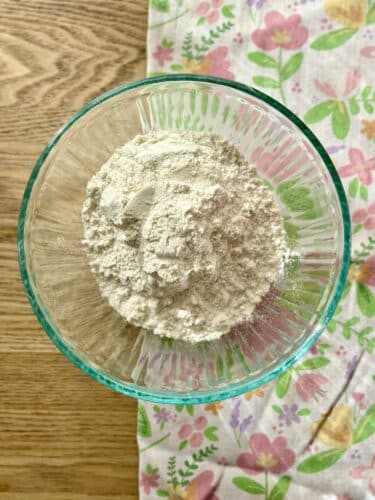 A bowl filled with all-purpose flour, sitting on a wooden surface, surrounded by a floral-patterned fabric.