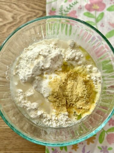 A mixing bowl containing flour, olive oil, and nutritional yeast, with a light floral-patterned cloth in the background.