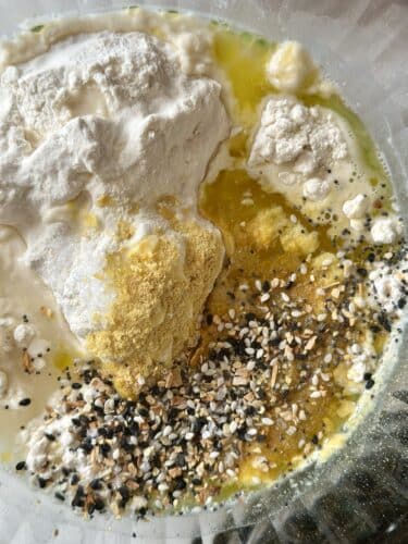 A close-up view of a mixing bowl containing flour, olive oil, everything bagel seasoning, nutritional yeast, and other dry ingredients, ready to be combined into dough.