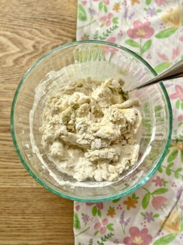 A mixing bowl containing a coarse mixture of flour and water, with a spoon resting inside, on a floral-patterned cloth.