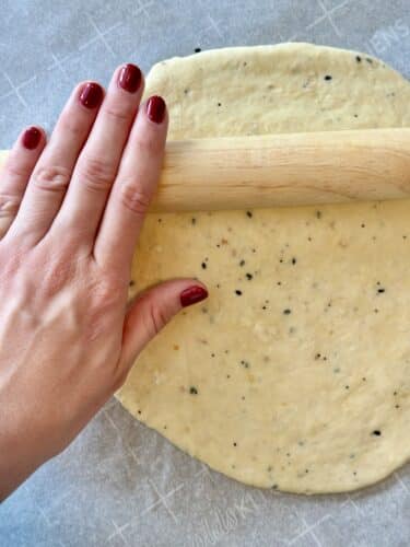 A hand with red nails using a rolling pin to flatten a piece of dough speckled with seasoning on a parchment-lined surface.