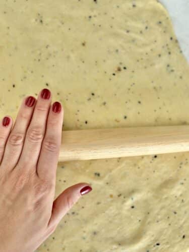 A hand with red nail polish rolling out dough for homemade crackers, with visible flecks of seasoning on a light-colored surface.