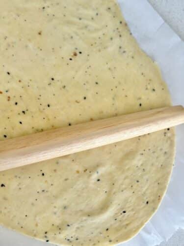 A rolling pin on a sheet of dough seasoned with everything bagel spices, spread thin on parchment paper.