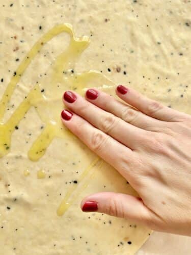 A hand brushing olive oil over rolled-out cracker dough, with visible seasoning flecks.