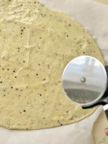 A round, flattened dough for homemade everything bagel crackers on parchment paper, with a pizza cutter placed nearby.