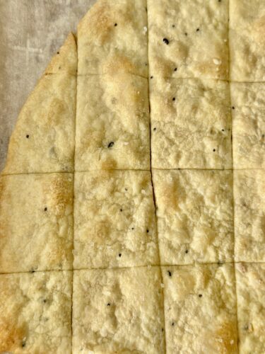Close-up view of freshly baked homemade everything bagel crackers, scored and ready to break into pieces, showing a golden-brown surface with seasoning flecks.