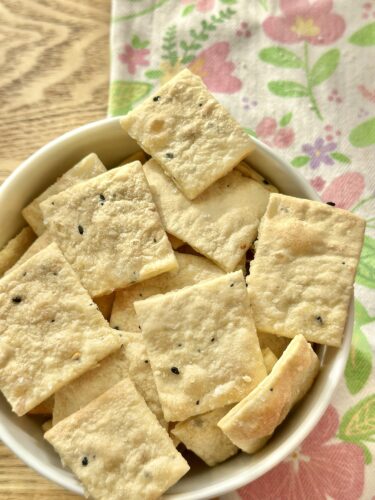 A bowl filled with homemade everything bagel crackers, cut into square shapes, resting on a table with a floral cloth in the background.