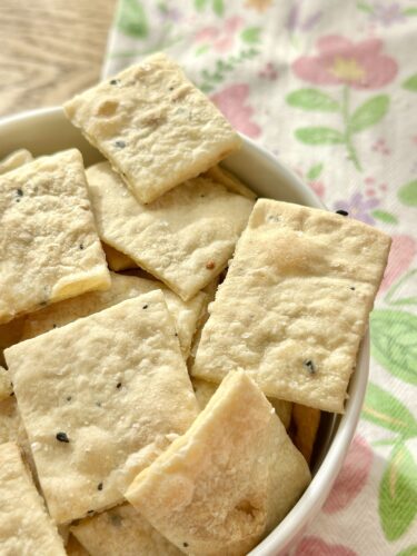 A bowl filled with homemade everything bagel crackers, showcasing a variety of crisp, rectangular shapes, resting on a patterned cloth.