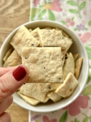 A hand holding a square homemade cracker above a bowl filled with more crackers, showcasing their crispy texture.