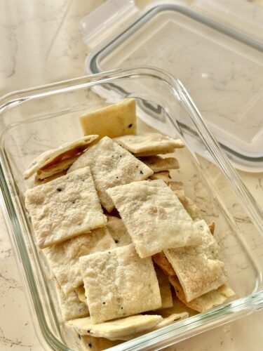 A glass storage container filled with homemade crackers, showcasing a variety of perfectly baked, crispy, and golden-brown pieces, some sprinkled with seasoning.