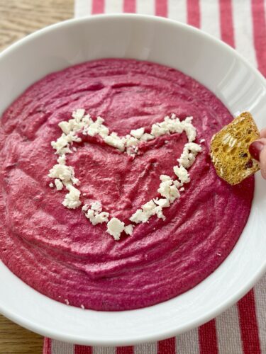 A vibrant, pink whipped beet dip garnished with crumbled feta in the shape of a heart, served in a white bowl, with a piece of bread being dipped into it. The bowl is placed on a striped tablecloth.
