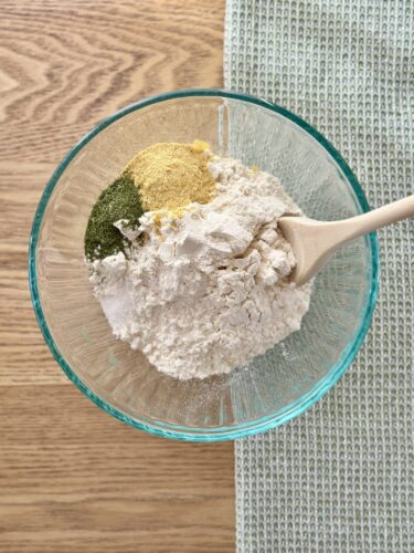 A glass bowl filled with flour and various spices, including dill and nutritional yeast, with a wooden spoon resting inside.