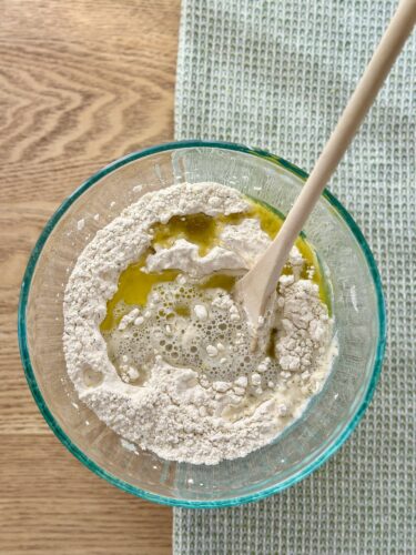 A mixing bowl containing flour, olive oil, and water, with a wooden spoon stirring the ingredients together, placed on a wooden surface.