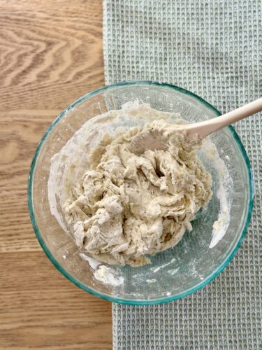 A glass bowl containing a mixture of dough for homemade dill crackers, with a wooden spoon resting inside, placed on a textured gray surface.