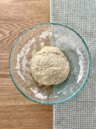 A glass bowl containing a ball of dough, resting on a wooden surface with a textured gray cloth beside it.