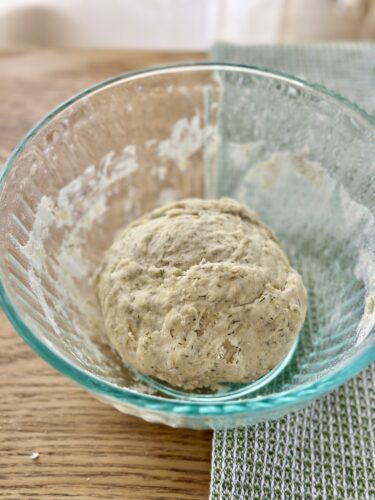 A glass bowl containing a ball of dough made with herbs, resting on a wooden surface next to a green fabric.