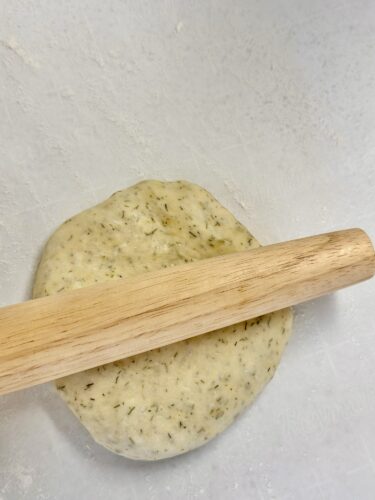 A wooden rolling pin resting on a round, herb-infused dough on a floured surface, ready to be rolled out.