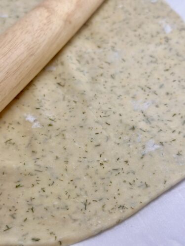 Close-up of rolled-out cracker dough sprinkled with dill, with a wooden rolling pin partially visible.