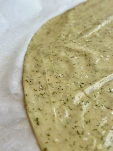A close-up of rolled-out cracker dough seasoned with herbs and sprinkled with sea salt on parchment paper.