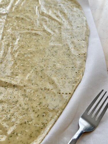 A rolled-out cracker dough sprinkled with herbs, resting on parchment paper with a fork beside it.