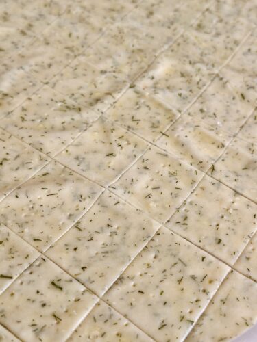A close-up view of rolled cracker dough, seasoned with dill and cut into squares, ready to be baked.